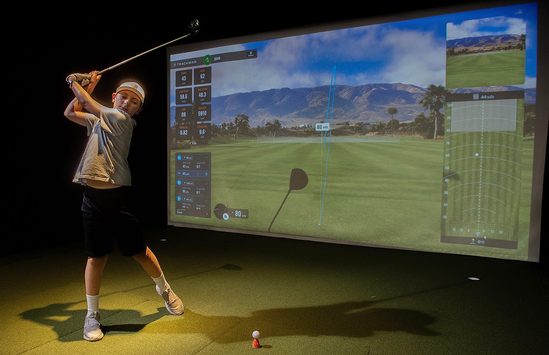A young golfer swings in a Trackman simulator as metrics from his previous swing flash on the screen in front of him. 